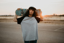Stylish Hipster Teenage Guy Holding Skateboard On Behind Head And Looking At Camera On Empty Road In Sunset