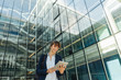 © Hernandez And Sorokina/ADDICTIVE STOCK - Low angle of cheerful entrepreneur smiling and browsing tablet while standing outside building with glass walls in downtown