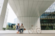 © Hernandez And Sorokina/ADDICTIVE STOCK - Delighted businesspeople smiling and browsing laptop together while sitting outside modern building near bicycle on city street