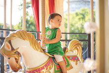 Pensive Curious Child In Green Casual Clothes Looking Away While Riding Fabulous Carnival Horse Alone In Amusement Park In Sunny Day