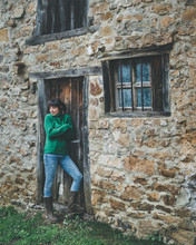 Casual Female In Green Sweater And Jeans Standing With Crossed Arms Next To Door Of Old Stone Country House And Feeling Cold On Gloomy Damp Autumn Day