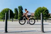 Side View Of Athletic Male In Sunglasses Wearing White Shirt And Black Shorts Riding Bike On City Roadway With Green Trees On Roadside On Summer Day With Blue Sky