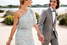 Pleased Young Groom In Wedding Suit Proudly Looking At Beautiful Blonde Haired Bride In Stylish Dress Behind At Empty Sandy Seashore