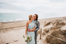 Pleased Young Groom In Wedding Suit Hugging And Kissing Blonde Haired Bride In Stylish Dress Behind At Empty Sandy Seashore