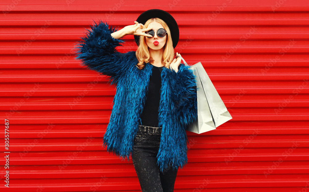 Attractive woman with shopping bags, stylish female model showing peace gesture wearing blue faux fur coat, black round hat and sunglasses over red wall background - obrazy, fototapety, plakaty 