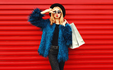 Attractive Woman With Shopping Bags, Stylish Female Model Showing Peace Gesture Wearing Blue Faux Fur Coat, Black Round Hat And Sunglasses Over Red Wall Background