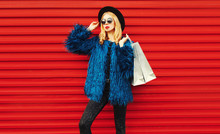 Attractive Woman Posing With Shopping Bags, Stylish Female Model Wearing Blue Faux Fur Coat, Black Round Hat And Sunglasses Over Red Wall Background