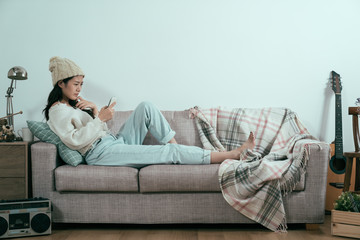 Wall Mural - isolated white background. Lonely sad asian korean girl sitting alone in house on sofa in morning and looking at smart phone waiting for phone call. lady in warm sweater and bobble hat for winter