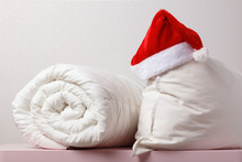 Close-up Of A Rolled Duvet And A Pillow With A Cap Of Santa Claus Lying On The Chest, On The Background Light Walls