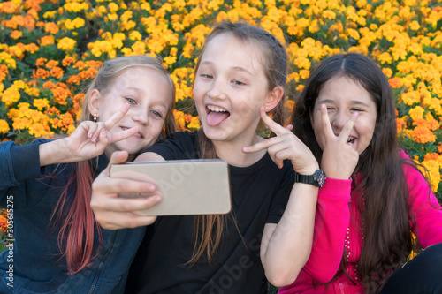 three teen girls take a selfie on Phone on a sunny day and laugh. Girls are showing victory sign with their fingers.
