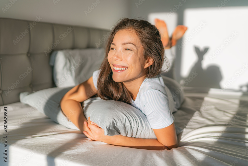 Bed woman waking up early morning happy enjoying sun on comfortable mattress and pillow. Asian girl relaxing in bedroom smiling. Natural beauty after healthy good night sleep sleeping in on weekend. - obrazy, fototapety, plakaty 