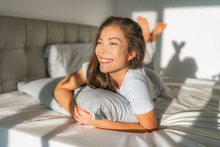 Bed Woman Waking Up Early Morning Happy Enjoying Sun On Comfortable Mattress And Pillow. Asian Girl Relaxing In Bedroom Smiling. Natural Beauty After Healthy Good Night Sleep Sleeping In On Weekend.