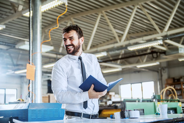 Handsome smiling caucasian bearded graphic engineer holding notebook while standing in printing shop. In background are printing machines.