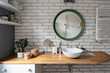Modern bathroom interior in industrial style with white bricky wall. Wooden countertop, stylish washinbasin and round mirror in bright indoors.