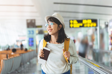 happy asian woman wear glasses, hat with yellow backpack is holding flying ticket, passport while wa