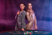 Two Girls Are Playing Poker At Casino, Holding Glasses Of Champagne, Posing At A Table With Chips, Cards On It. Black, Smoke Background. Close-up.