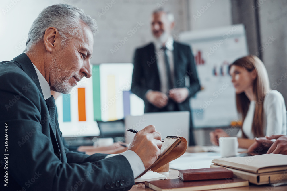Mature businessman writing something down and smiling while working together with colleagues in the board room - obrazy, fototapety, plakaty 