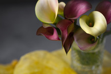 Bunch Of Red And Yellow Calla Lilies In A Clear Vase; Yellow Fabric In Background; Gray Textured Background