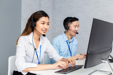 Cheerful Asian Man And Woman In Headsets Smiling And Typing On Computer Keyboard While Working In Office Of Call Center