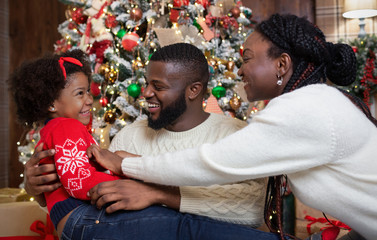 Wall Mural - Joyful black parents tickling their little daughter, celebrating Christmas together