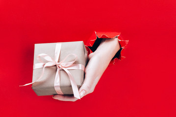 Female hand holding gift box wrapped in craft paper with pink ribbon through a hole in red background.