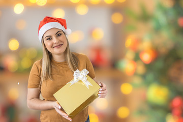 Young woman smiling wearing Christmas hat, holding a golden gift box on blur background