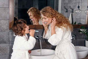 Attractive smiling young mother in dressing gown cleans her teeth together with handsome little son near washbasin with mirror. Happy family concept.