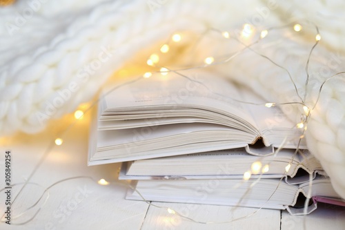 Winter books. Reading in the winter holidays. White cover books , shining garland and white knitted scarf on a white wooden table. Christmas books. Winter reading. White book background.
