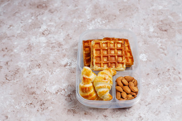 Delicious breakfast with almonds,red apples slices, waffles,croissants on plastic lunch box on light background