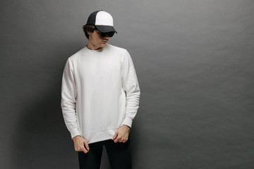 Wall Mural - Man wearing blank white sweatshirt and empty baseball cap standing over gray background. Sweatshirt or hoodie for mock up, logo designs or design print with free space.