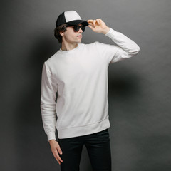 Wall Mural - Man wearing blank white sweatshirt and empty baseball cap standing over gray background. Sweatshirt or hoodie for mock up, logo designs or design print with free space.