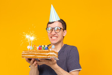 Wall Mural - Positive funny young asian guy with a cap and a burning candle and a homemade cake in his hands posing on a yellow background. Anniversary and birthday concept.