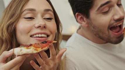 Sticker - Attractive happy couple man and woman lying on bed at home and enjoy eating tasty pizza 