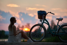 Beautiful Girl Sits Near A Vintage Bicycle With A Basket On A Blurry Background Of The Evening Sky At Sunset With A Fiery Sun. Girl In A Light Summer Dress And Sneakers Resting In Nature