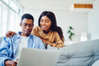 © BullRun - Smiling african american woman looking at freelance job of husband sitting with laptop computer at sofa in living room, positive dark skinned couple in love making shopping together  in web store.