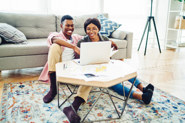 Cheerful african american hipster couple watching online movies on laptop computer resting in living room together, young marriage making video call via application on netbook at cozy apartment