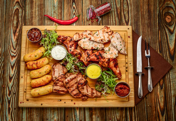 Wall Mural - Assorted delicious grilled meat and vegetables with fresh salad and bbq sauce on cutting board on wooden background. Big set of Hot Meat Dishes