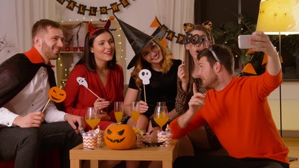 Sticker - friendship, holiday and people concept - group of happy smiling friends in halloween costumes of vampire, devil, witch and leopard taking selfie by smartphone at home party at night