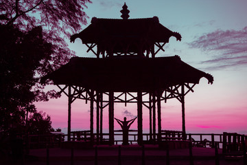 Backlit photo of young woman doing V-leg stretching on the table located at Vista China lookout, in Tijuca Forest in Rio de Janeiro. Colorful sky with shades of orange and blue and some clouds.