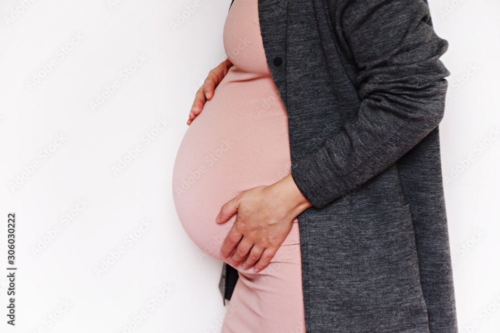 Beautiful pregnant woman touching her belly with hands on a white background. Young mother anticipation of the baby. Image of pregnancy and maternity. Close-up, copy space, indoors. - obrazy, fototapety, plakaty 
