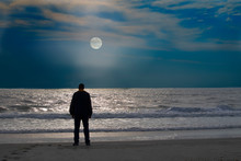 A Man Stands Alone On At The Edge Of The  Calm Ocean  Looking At Rising Moon