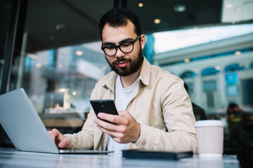 Asian businessman holding smartphone checking notifications about incoming mail while working remotely on laptop computer in coffee shop, hipster guy in eyewear using technologies for learning online.