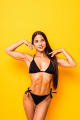 Wall Mural - Young sexy slim tanned woman in black swimsuit posing against yellow background.