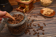 The Girl's Hand Takes A Wooden Spoon Of Coffee Beans From A Glass Jar, A Coffee Grinder, Pieces Of Brown Sugar On A Wooden Background. Close Up.