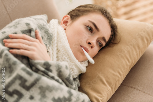 Cuadros en Lienzo ill woman lying on sofa under blanket and measuring temperature