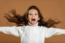 Happy Funny Little Child Girl In White Sweater Showing Her Tongue On Beige Background. Facial Expression