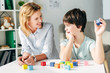 © LIGHTFIELD STUDIOS - Smiling child psychologist looking at kid with dyslexia and sitting at table with building blocks