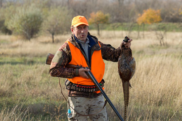 Wall Mural - A man with a gun in his hands and an orange vest on a pheasant hunt in a wooded area in cloudy weather. A hunter with a pheasant in his hands.