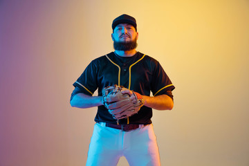 Baseball player, pitcher in a black uniform practicing and training on gradient background in neon light. Young professional sportsman in action and motion. Healthy lifestyle, sport, movement concept.