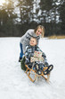 © sofiko14 - Beautiful family of young mother and son enjoying snowy winter day outdoors having fun sledging. Boy is sitting on the wooden decorated sledge with presents, while mom is pushing him
